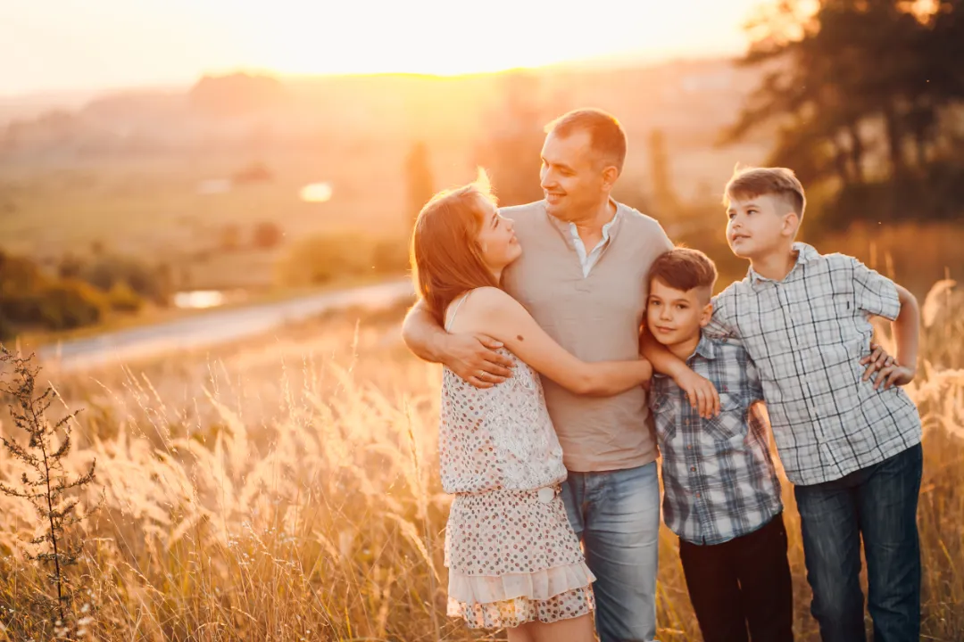 A family of four stands together in a sunlit field, smiling and embracing, with golden grass and a picturesque landscape behind them.